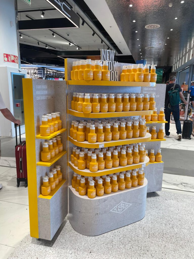 Expositor de productos Sol de Janeiro en el aeropuerto de Málaga, con estanterías amarillas llenas de botellas, diseño retail tropical y montaje experiencial.